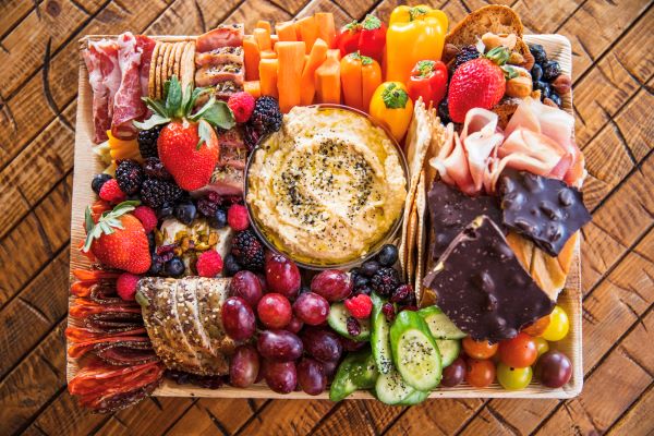 A wooden platter with hummus in the center, surrounded by an assortment of fruits, vegetables, meats, cheeses, and crackers, embodies the essence of healthy food franchises.