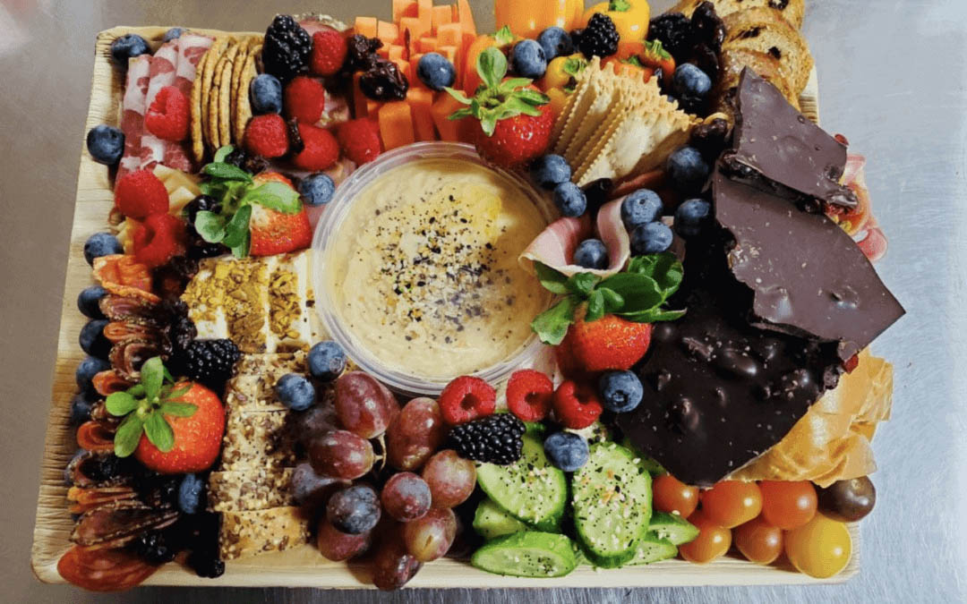 A platter with a central bowl of dip surrounded by strawberries, grapes, blueberries, carrots, crackers, meats, cheese, and chocolate.