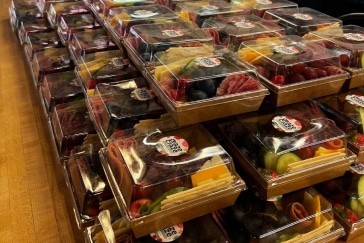 Stacks of packaged charcuterie boxes with assorted meats, cheeses, and fruits arranged neatly on a wooden surface.