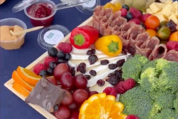 A platter with assorted fruits, vegetables, cheese, meats, chocolate, and dips. Includes grapes, berries, orange slices, peppers, broccoli, brie, and salami rolls.