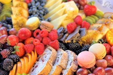 A colorful array of sliced bread, macarons, assorted fruits, cheese slices, and berries.