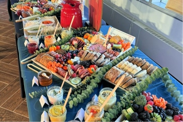 A food spread with various cheeses, breads, fruits, meats, dips, and pastries are arranged on a table with greenery and small labels. A red pitcher is in the center.