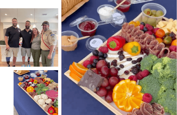 A group of four people stands indoors. Adjacent is a table with a charcuterie board featuring an assortment of meats, cheeses, fruits, and vegetables.